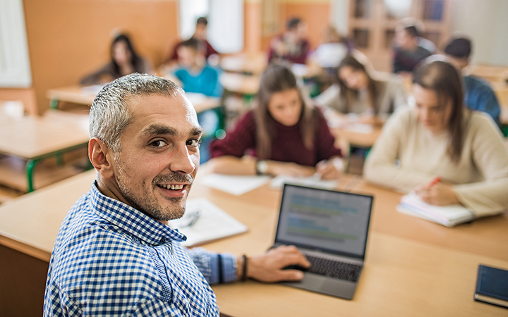 Smiling male teacher on laptop in front of classroom